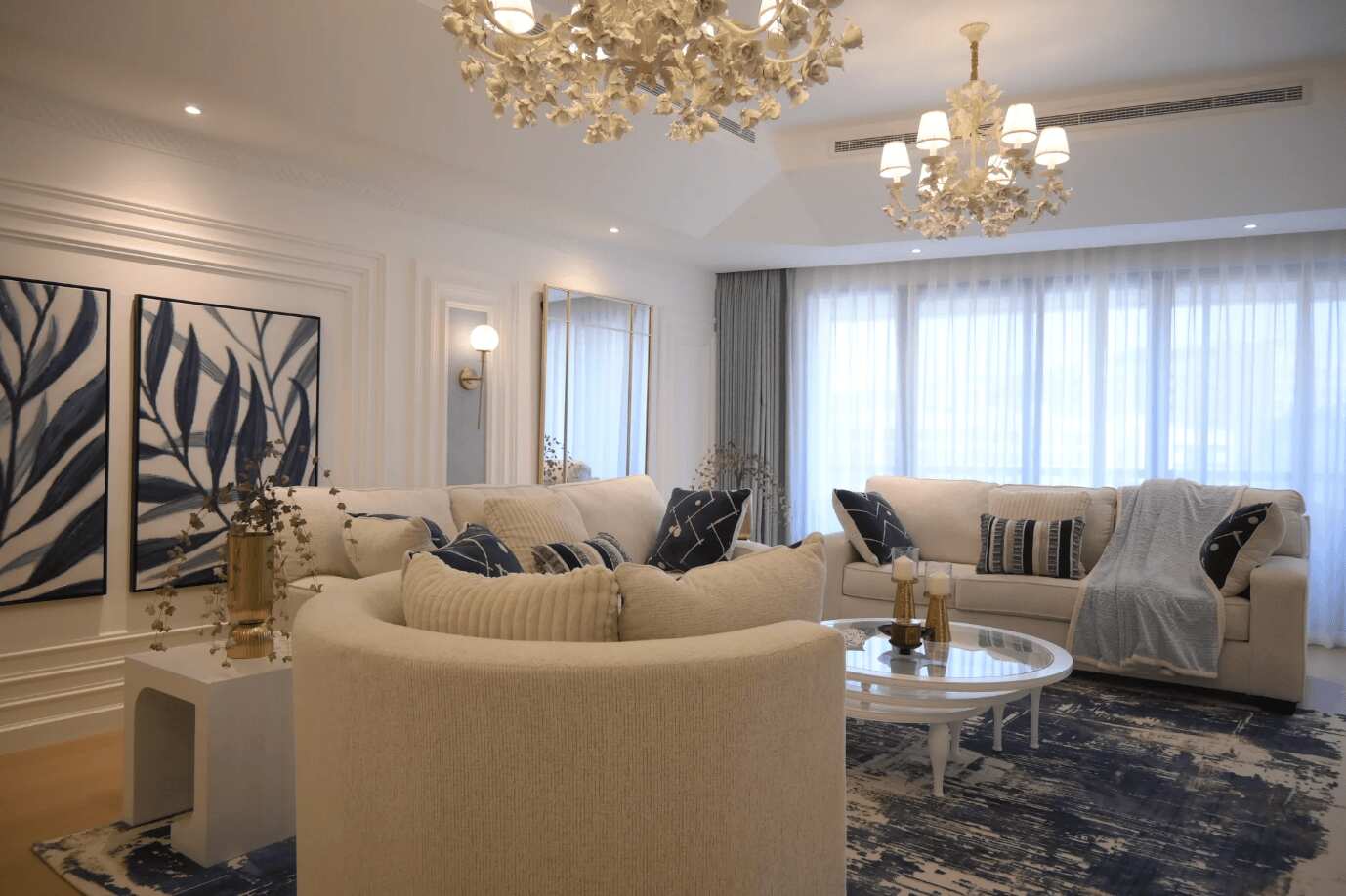 This elegant living room features neutral beige sofas, blue and white accents, and a luxurious chandelier, with modern artwork on the walls.