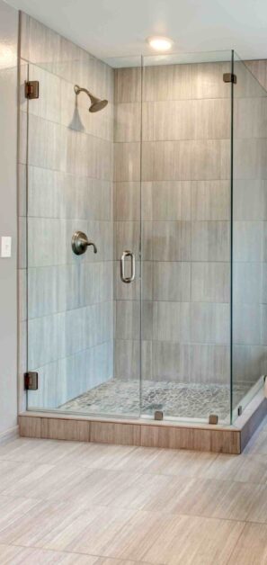 Modern walk-in glass shower with light gray tiled walls and a pebble stone floor, featuring a brushed nickel showerhead and handle.