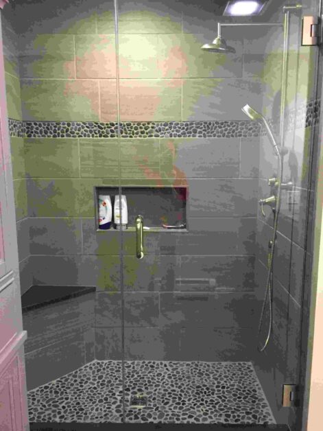 Modern glass-enclosed shower with dark gray tiles, pebble stone floor and showerheads including a handheld sprayer.