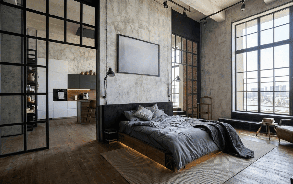 The main focal point in this minimalist bedroom design by loakdesigns is the raw and earthy stone wall that flows up from the bed to the ceiling with a hidden light washing the textures. Earthy stone textures are highly practical for homes in the UAE because these materials naturally stay cool against the ambient heat. Because of this texture, you can do away with any kind of extra decor or artwork. A minimalist bed sits on top of a tactile rug that contrasts beautifully against the dark wooden floor and walls. The mid-height plants keep the corners full, and the minimalist bedroom curtains' heads are beautifully tucked inside the disconnected space of the ceiling to the walls. The overhead light provides a soft glow as evening falls, creating a luxurious and earthy space. 11. Industrial Style Minimalist Bedroom Design Idea
